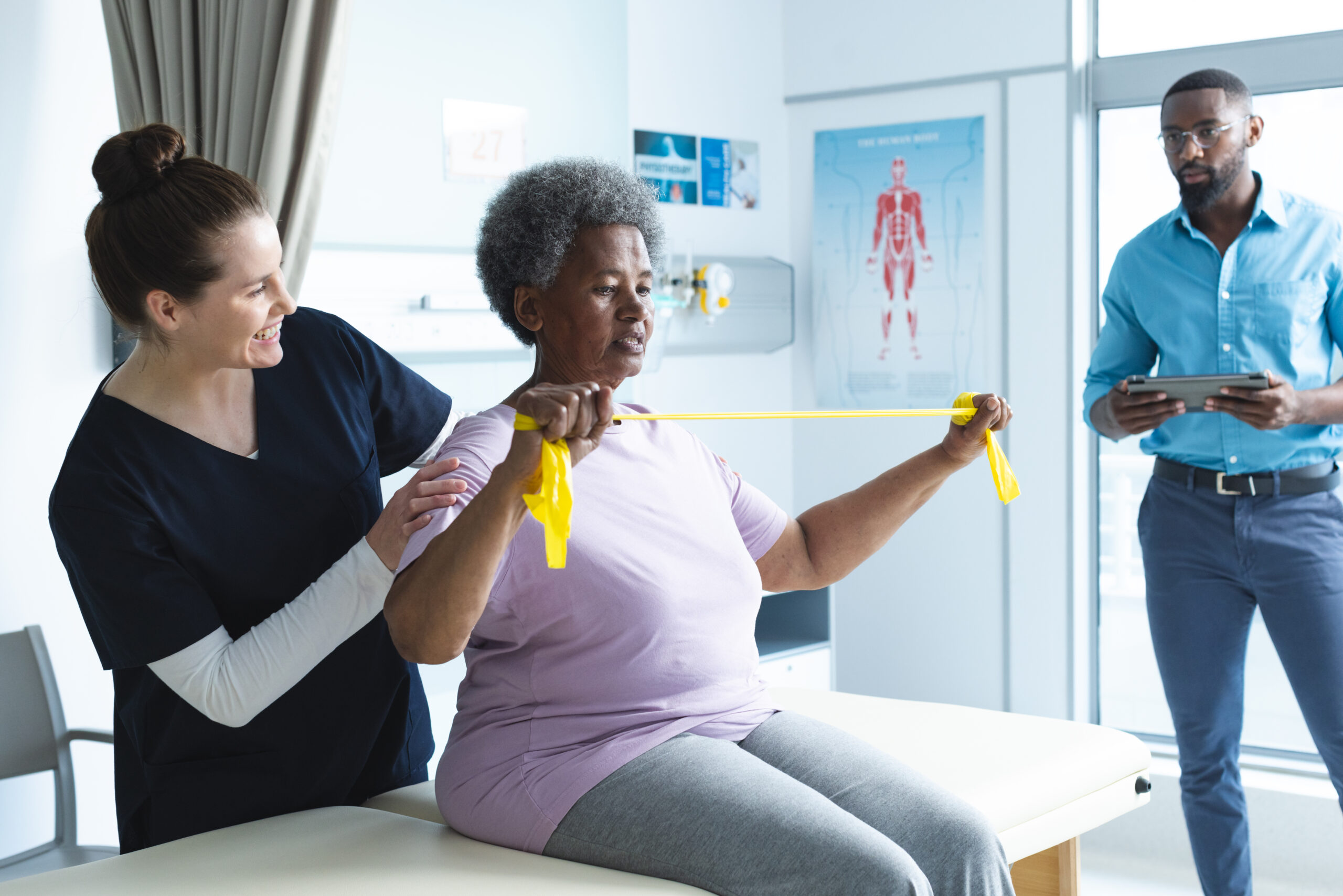 Diverse senior female patient exercising with band and female doctor advising in hospital room. Medicine, healthcare and medical services, unaltered.