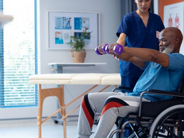 Diverse female physiotherapist helping senior male patient in wheelchair using dumbbells, copy space. Hospital, medical and healthcare services.
