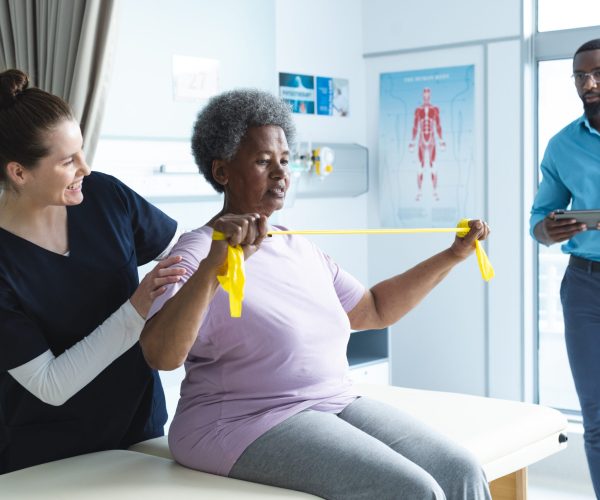 Diverse senior female patient exercising with band and female doctor advising in hospital room. Medicine, healthcare and medical services, unaltered.
